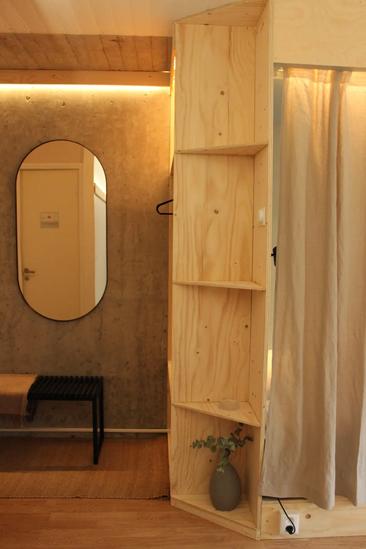 A warmly lit hotel room interior with plywood built-in shelving, an oval mirror and fabric canopy details.