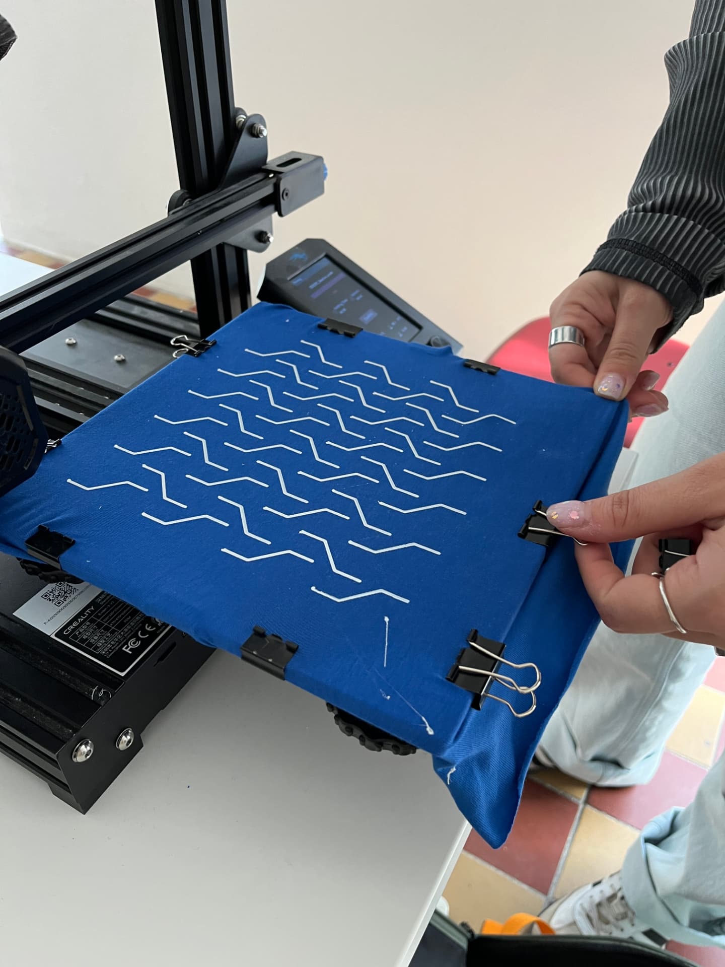 A desktop 3D printer printing white zigzag lines onto a stretched blue fabric held by binder clips.