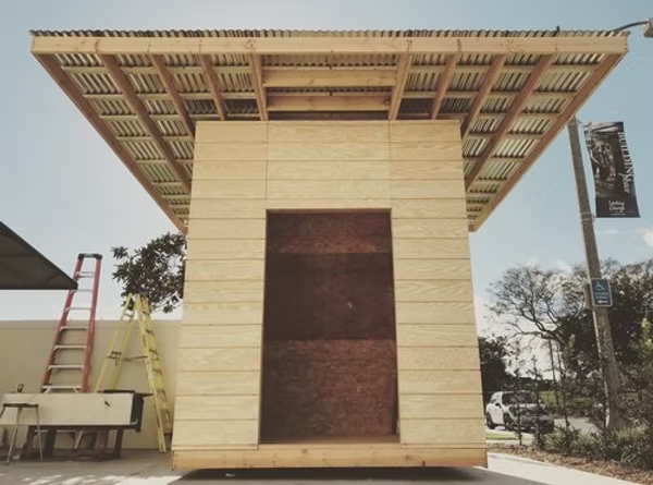 Compact wooden gallery structure with a slatted roof and plywood walls installed outdoors.