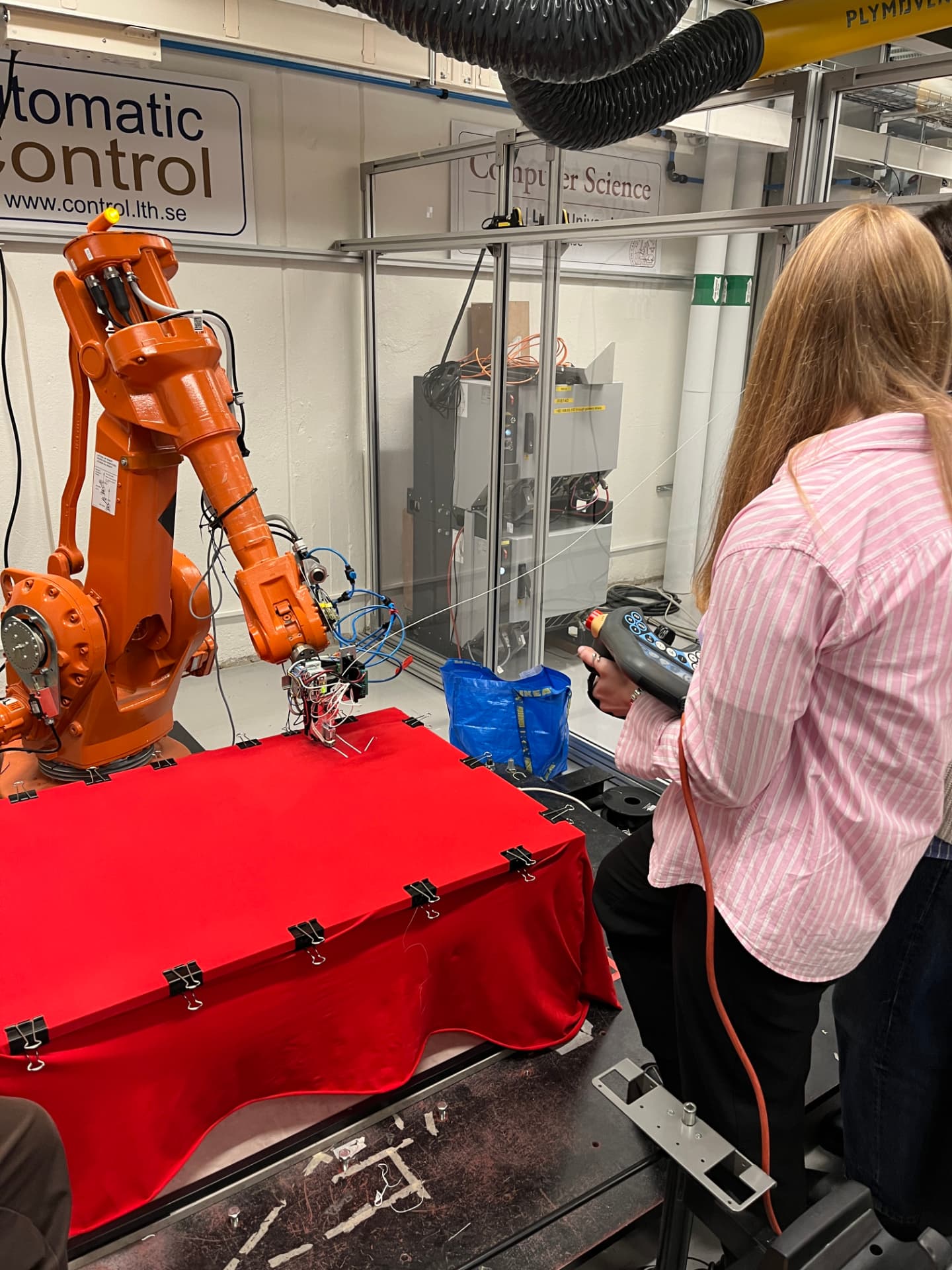 A person operates an orange ABB industrial robot arm with a custom extruder printing onto red tensioned fabric in a lab.