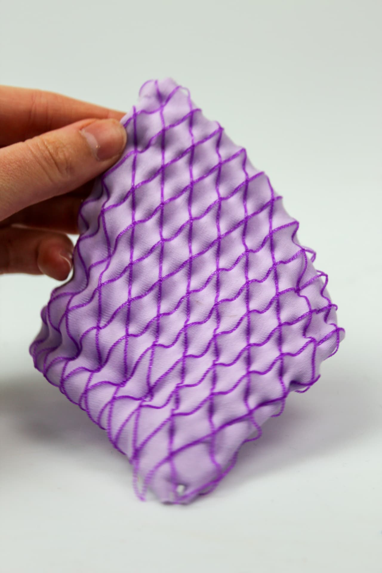 Hand holding a lilac textile sample with a purple printed diamond grid buckling into relief.