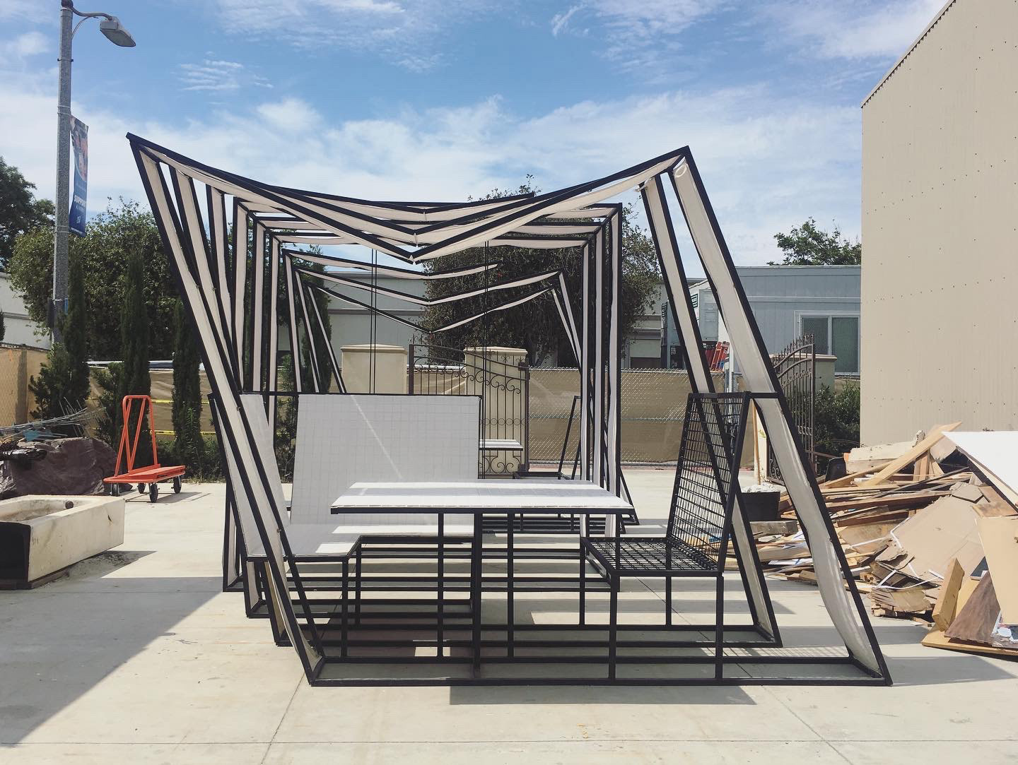 A black steel and white fabric pergola structure with built-in seating in an outdoor urban lot.